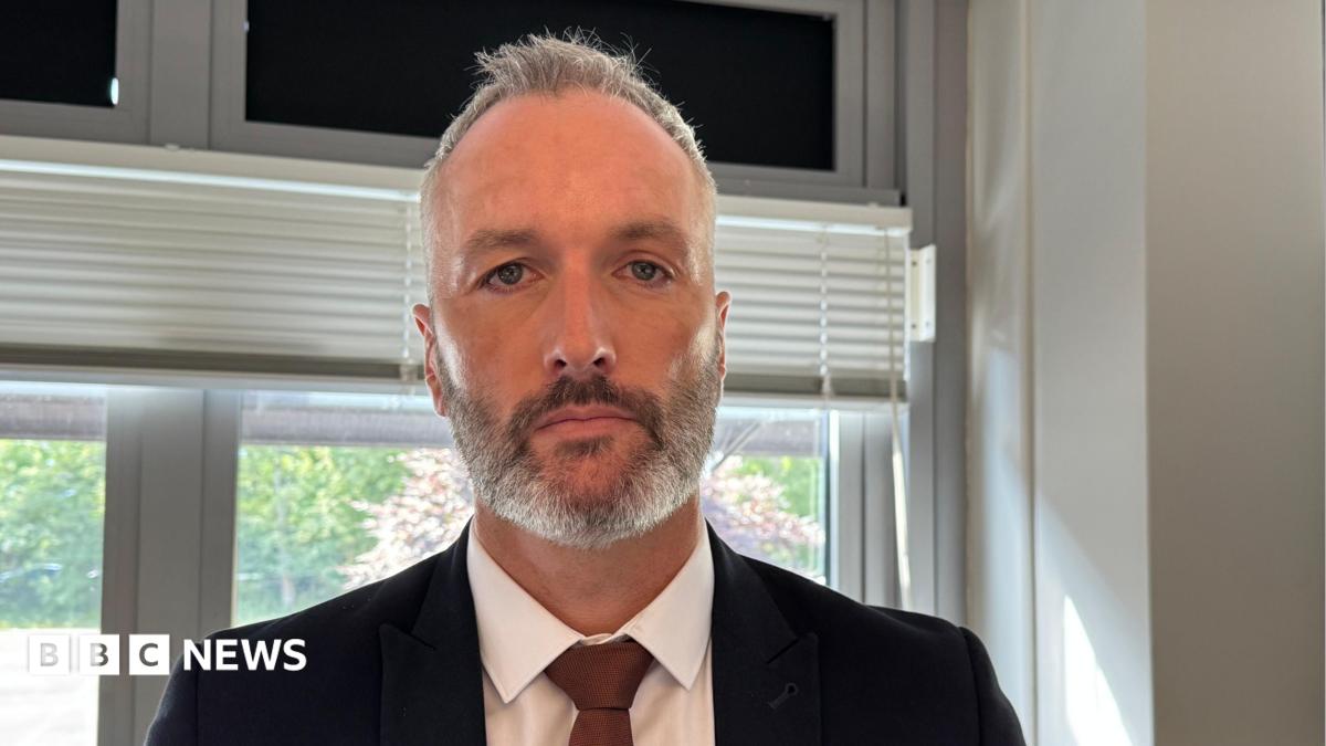 Det Con Lee Harshey-Jones. He looks at the camera with a neutral expression, and wears a white shirt, black suit jacket and bronze tie. He has short grey hair and short stubble.