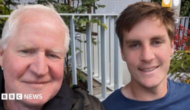 Father and son smiling at the camera. The dad is on the right and is wearing a black jacket and the son is on the right and is wearing a blue t-shirt. Behind them is a metal fence.