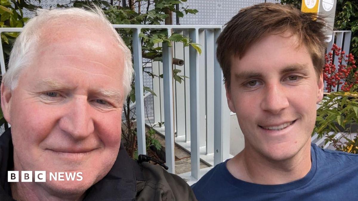 Father and son smiling at the camera. The dad is on the right and is wearing a black jacket and the son is on the right and is wearing a blue t-shirt. Behind them is a metal fence.