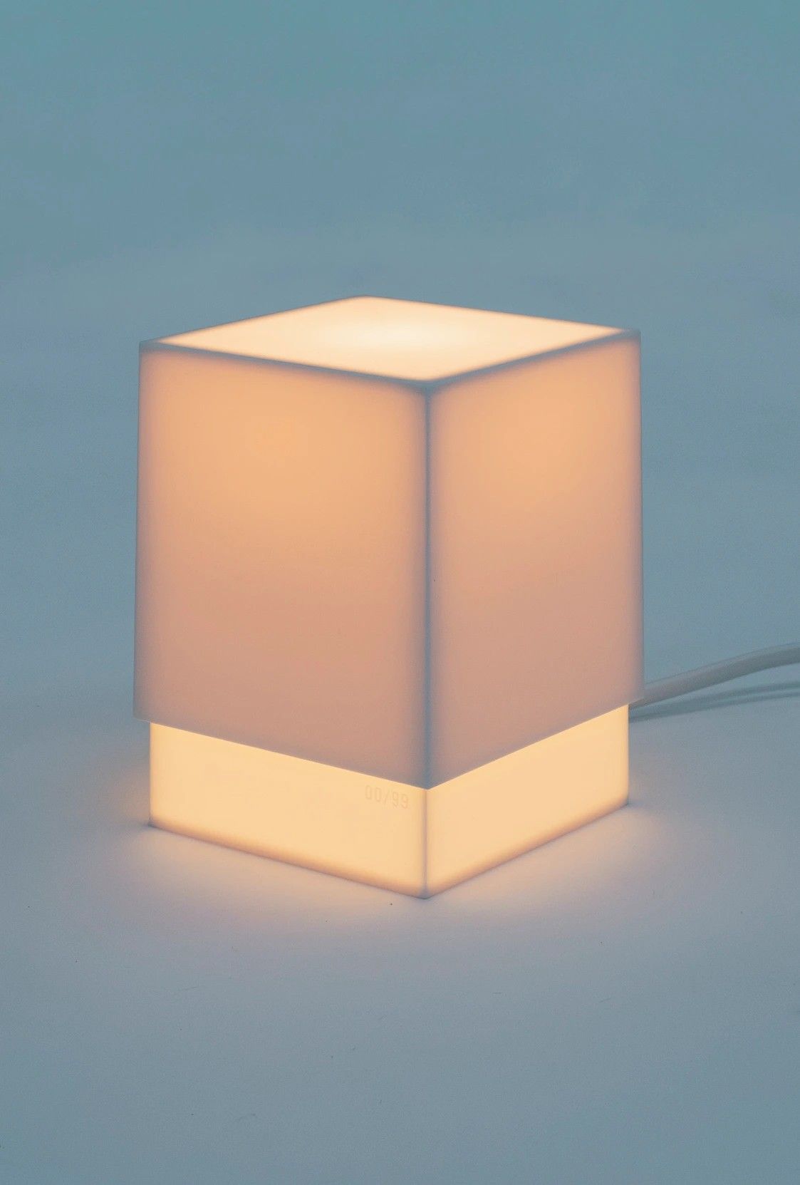 A square, box-like table lamp set against a pale blue background glows with pale pink light.
