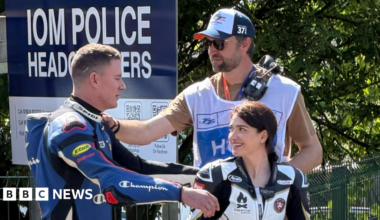 Channing Tatum on the left has blue and black leathers on, he's reaching for a hug to Eve Hewson on the right with black and white leathers on. She has dark hair tied up and is smiling at him. Director Reid Carolin is behind them with his hand on Channing's shoulder, wearing a TT bib over a t-shirt, with headphones over his shoulder, and wearing sunglasses and a TT cap.