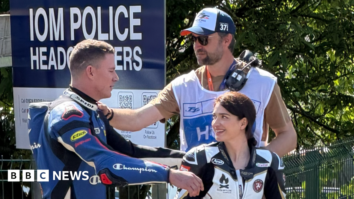 Channing Tatum on the left has blue and black leathers on, he's reaching for a hug to Eve Hewson on the right with black and white leathers on. She has dark hair tied up and is smiling at him. Director Reid Carolin is behind them with his hand on Channing's shoulder, wearing a TT bib over a t-shirt, with headphones over his shoulder, and wearing sunglasses and a TT cap.