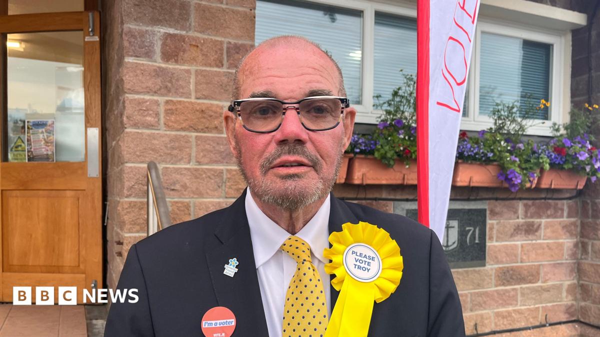 A man with very short grey hair, facial hair and glasses. He is wearing a white shirt, yellow tie with blue dots, and a dark blazer. He has a yellow rosette on his lapel, which says 'Please Vote Troy'. On the other side of his jacket is a sticker which says 'I'm a voter'. He is standing in front of a brick building with a wooden and glass door.