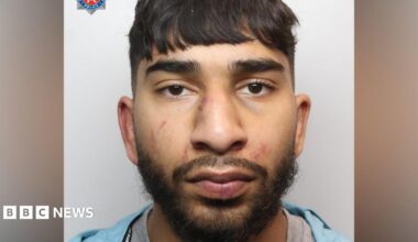 A mugshot of an Asian man in his early 20s, who has black hair cut into a fringe and black facial hair. He has red marks to his face  and is looking into the camera with a blank expression.