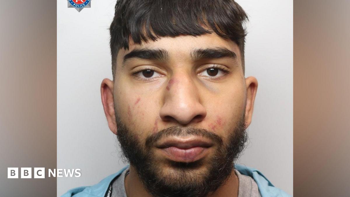 A mugshot of an Asian man in his early 20s, who has black hair cut into a fringe and black facial hair. He has red marks to his face  and is looking into the camera with a blank expression.