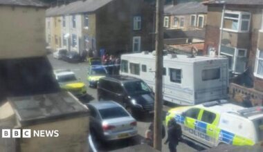 A number of police vehicles and officers stand behind a cordon in a street of terraced houses.
