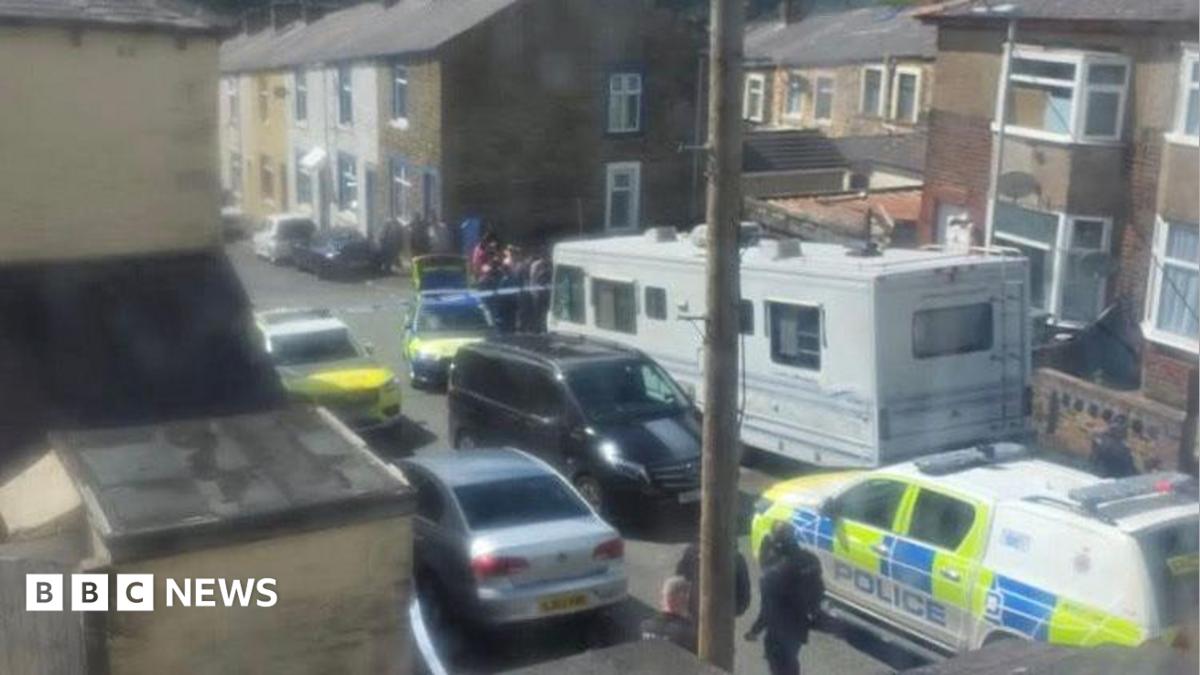 A number of police vehicles and officers stand behind a cordon in a street of terraced houses.