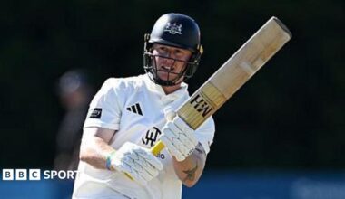Miles Hammond pulls the ball away through mid-wicket. He is holding his bat high to the right of the picture. He is wearing his whites and has a dak blue helmet on