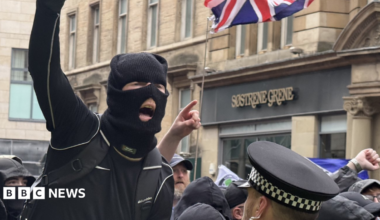 A masked individual in dark clothing raises a clenched fist while standing close to a police officer in a high-visibility jacket and checkered hat, with a crowd gathered behind and a Union Jack flag visible above on a city street lined with stone buildings and shopfront signage reading “Sostrene Grene.”