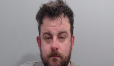A man looks into the camera. He has a dark beard and dark, dishevelled hair. He is wearing a grey, round neck top and is standing against a grey background.