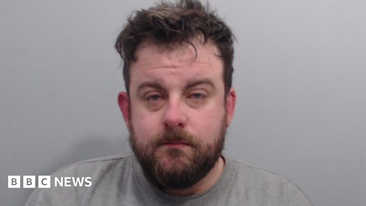 A man looks into the camera. He has a dark beard and dark, dishevelled hair. He is wearing a grey, round neck top and is standing against a grey background.