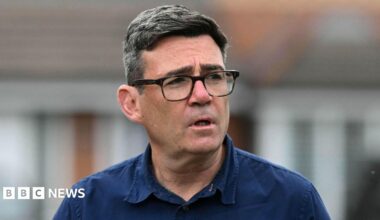Andy Burnham photographed from the shoulders up. He is wearing a navy blue shirt and black glasses. He is not looking at the camera.