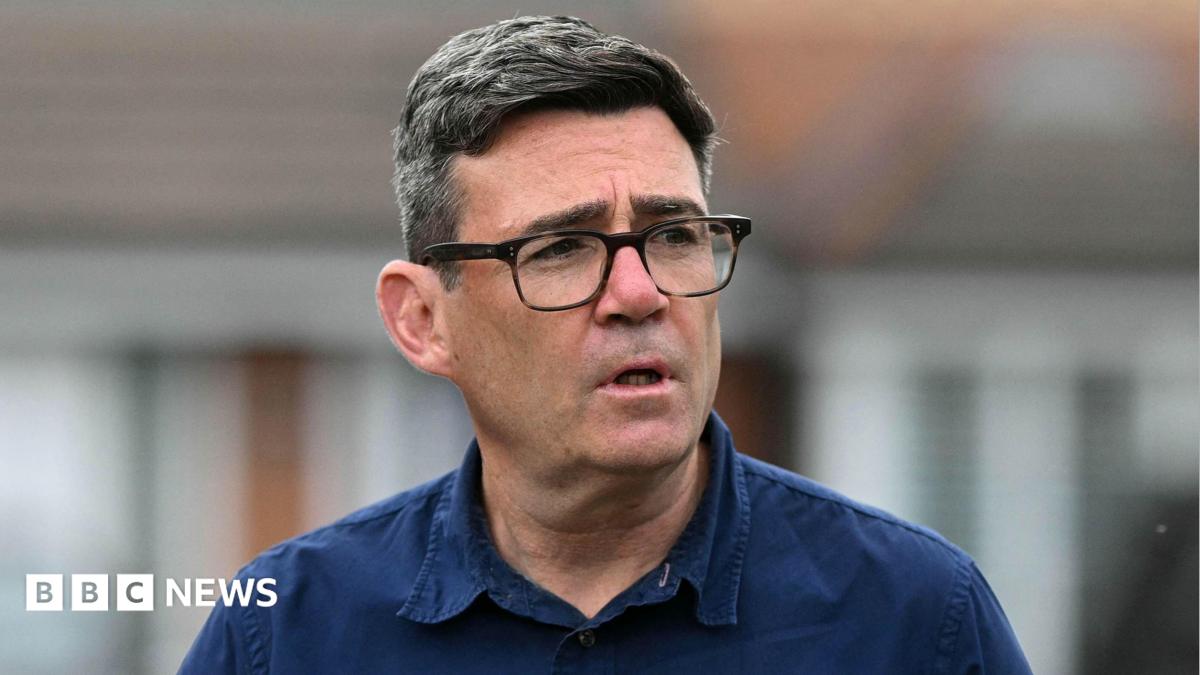 Andy Burnham photographed from the shoulders up. He is wearing a navy blue shirt and black glasses. He is not looking at the camera.