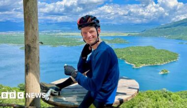 Tom Williamson in cycling clothes and a bike helmet, sitting on a wooden picnic table and smiling. A beautiful landscape with a lake, green islands and mountains is visible behind him.