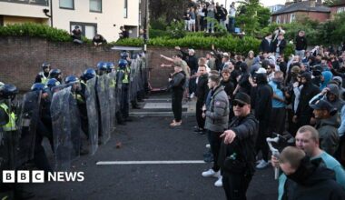 A line of riot police face off against a large group of protestors in the middle of a road.