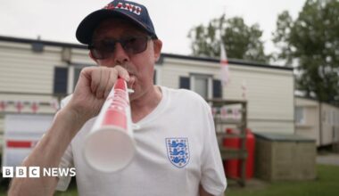 Richard blows an England themed vuvuzela trumpet, wearing a blue England Cap, white England shirt and sunglasses. Behind him is his static caravan decorated with England flag bunting.