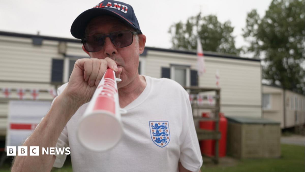 Richard blows an England themed vuvuzela trumpet, wearing a blue England Cap, white England shirt and sunglasses. Behind him is his static caravan decorated with England flag bunting.
