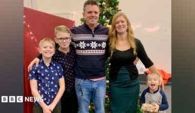 A family of five stands together in front of a Christmas tree in a group hug. On the left side, under their father's arm, are two blonde boys. In the centre is a man with short grey hair wearing a navy blue Christmas jumper, to his right is a woman with long strawberry blonde hair wearing a black top and green skirt, and to her right is a small boy with blonde hair and an excited expression.