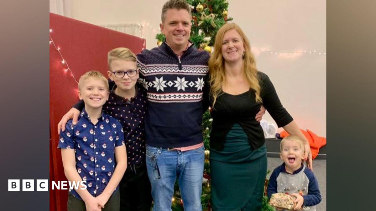A family of five stands together in front of a Christmas tree in a group hug. On the left side, under their father's arm, are two blonde boys. In the centre is a man with short grey hair wearing a navy blue Christmas jumper, to his right is a woman with long strawberry blonde hair wearing a black top and green skirt, and to her right is a small boy with blonde hair and an excited expression.