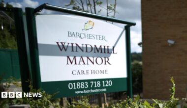 A white sign outside a building reading BARCHESTER WINDMILL MANOR CARE HOME 01883 718 120