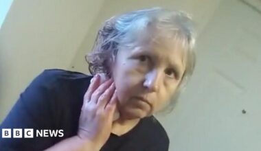 A screengrab of police body worn video showing Stefania Glowka stood in a corridor holding a bandage or tissue to a wound on her neck. She is a woman in her 60s with grey hair and is wearing a dark top.