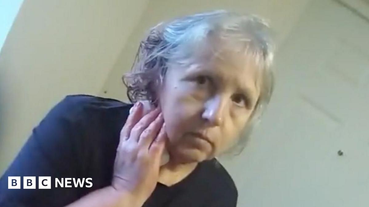 A screengrab of police body worn video showing Stefania Glowka stood in a corridor holding a bandage or tissue to a wound on her neck. She is a woman in her 60s with grey hair and is wearing a dark top.