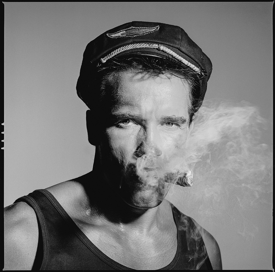 A square-format black-and-white portrait of Schwarzenegger in a Harley-Davidson cap and vest, staring directly into the camera while smoke billows from a cigar clamped between his lips.