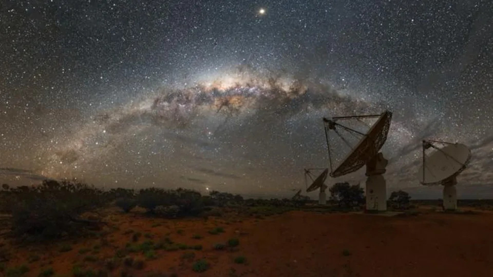 The Milky Way's heart glows beautifully in the sky. A bunch of radiowave detectors toward the right of the scene look up.