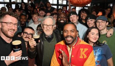 Film director Steven Spielberg (C) in a black leather jacket, green jumper and bandana around his neck, poses with fans inside a London pub while holding an half-full glass of Guinness. To his left is actor Colman Domingo in a red and yellow letterman jacket.