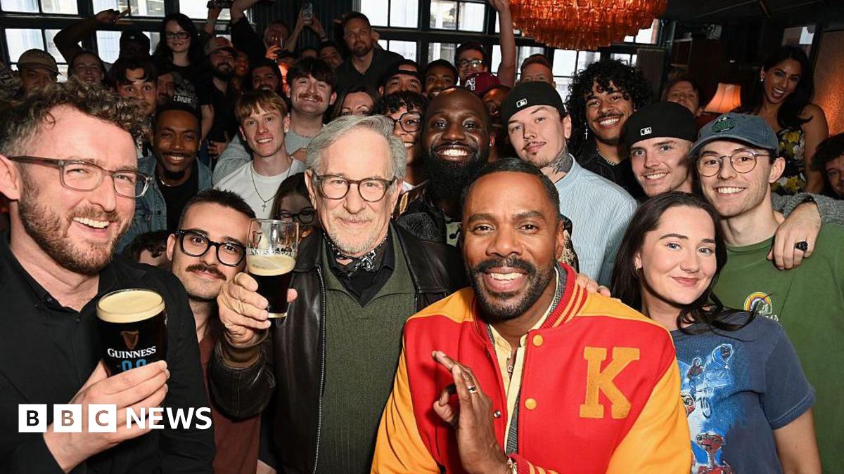 Film director Steven Spielberg (C) in a black leather jacket, green jumper and bandana around his neck, poses with fans inside a London pub while holding an half-full glass of Guinness. To his left is actor Colman Domingo in a red and yellow letterman jacket.