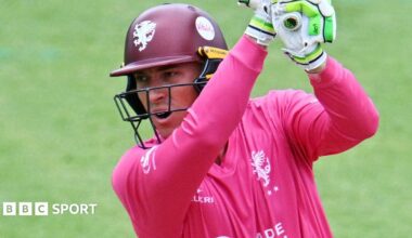 Tom Banton is playing a stroke looking over his right shoulder having just followed through his shot. He is wearing Somerset Sabres pink jersey and a navy blue helmet with the Somerset emblem of a Wyvern.