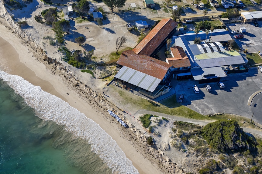 A drone view of ocean at an eroded beach, with a building sitting about 5 metres from a sand cliff.