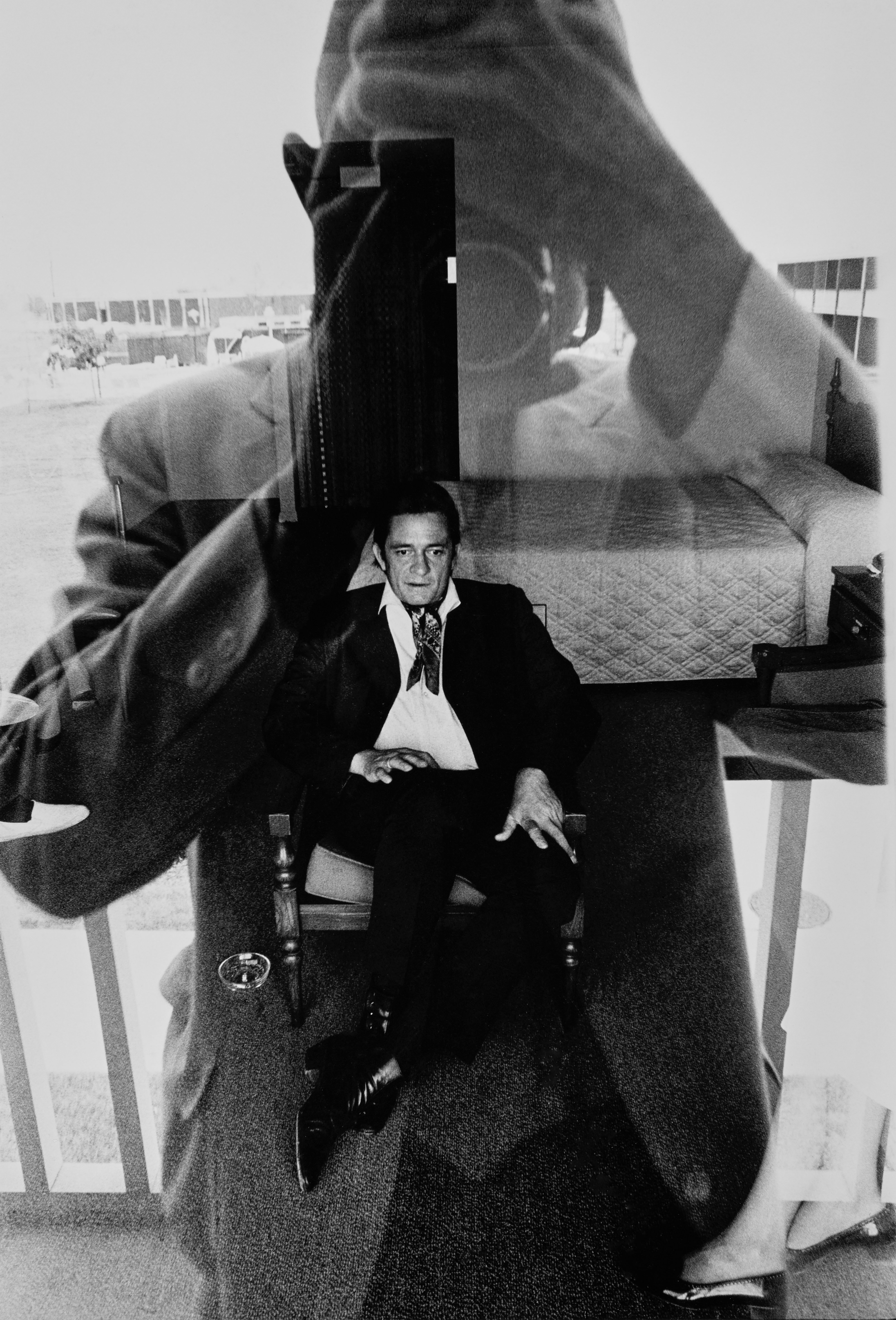 View, through the reflection of the photographer, of American musician Johnny Cash (1932 - 2003) as the latter sits in a chair in a motel room, November 1969. Cash wears a neckerchief, a white shirt, dark blazer, and trousers, and polished, buttoned boots. (Photo by Duane Michals/Conde Nast via Getty Images)