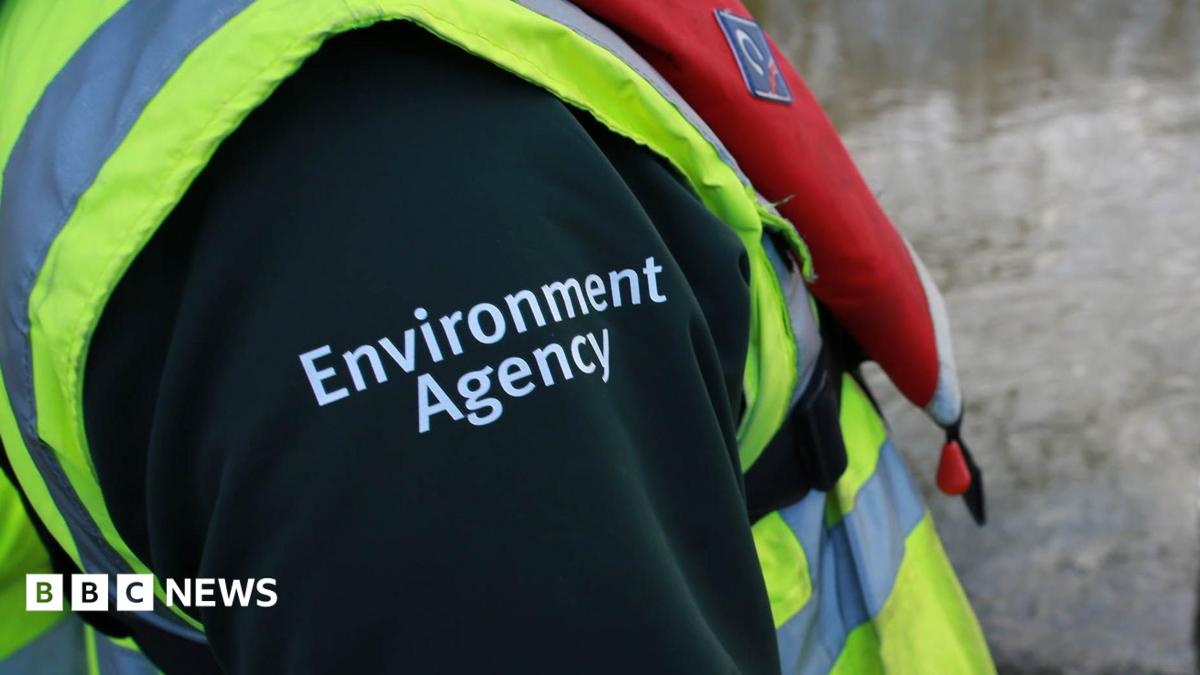 A close up of an Environment Agency employee's outfit. All that is visible is a high vis jacket and the shoulder of a black top with Environment Agency written on it in white.