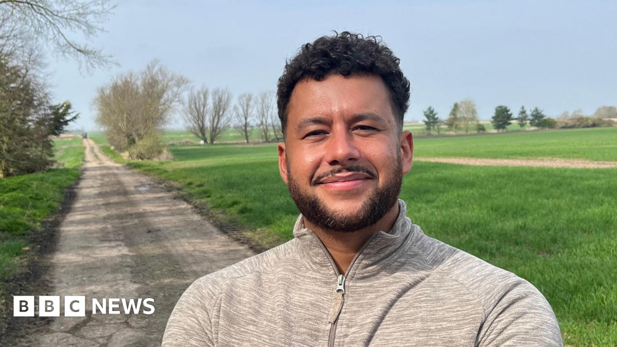 Ben Brooke has short, neat dark hair, a bit curly on top, he has a matching beard and moustache and is wearing a grey cotton sports top zipped up to his neck. Behind him are grass fields with a rough track wide enough for one vehicle running through it.