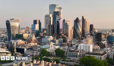 View of the major buildings of the City of London, headquarters to many financial firms, at dawn. The low sun off screen to the right illuminates the facades of the many multistorey skyscrapers, with the turrets of the Tower of London in the foreground.