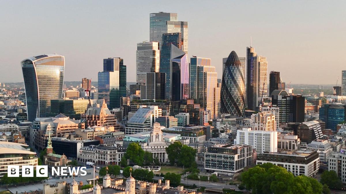 View of the major buildings of the City of London, headquarters to many financial firms, at dawn. The low sun off screen to the right illuminates the facades of the many multistorey skyscrapers, with the turrets of the Tower of London in the foreground.