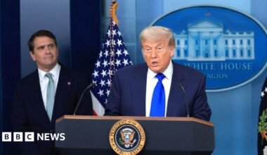 Trump is at the podium and speaking while wearing a dark blue suit and bright blue tie. To his right and behind him is Todd Blanche who is wearing a dark suit and indigo tie