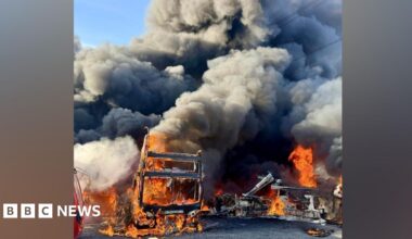A double-decker bus engulfed in flames, with smoke billowing from it.