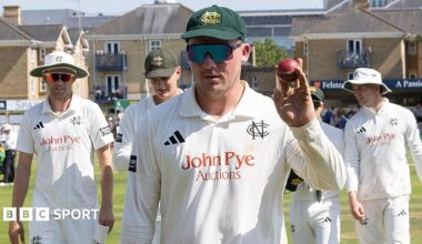 Liam Patterson-White holds up a red cricket ball with players surrounding him