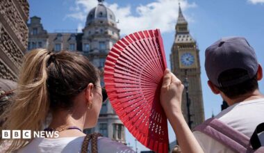 A close-up, over-the-shoulder shot from behind a woman with blonde hair in a ponytail holding up an open red hand fan. Next to her, a man wears a grey baseball cap. In the blurred background, London's Elizabeth clock tower rises against a clear blue sky on a bright, sunny day.