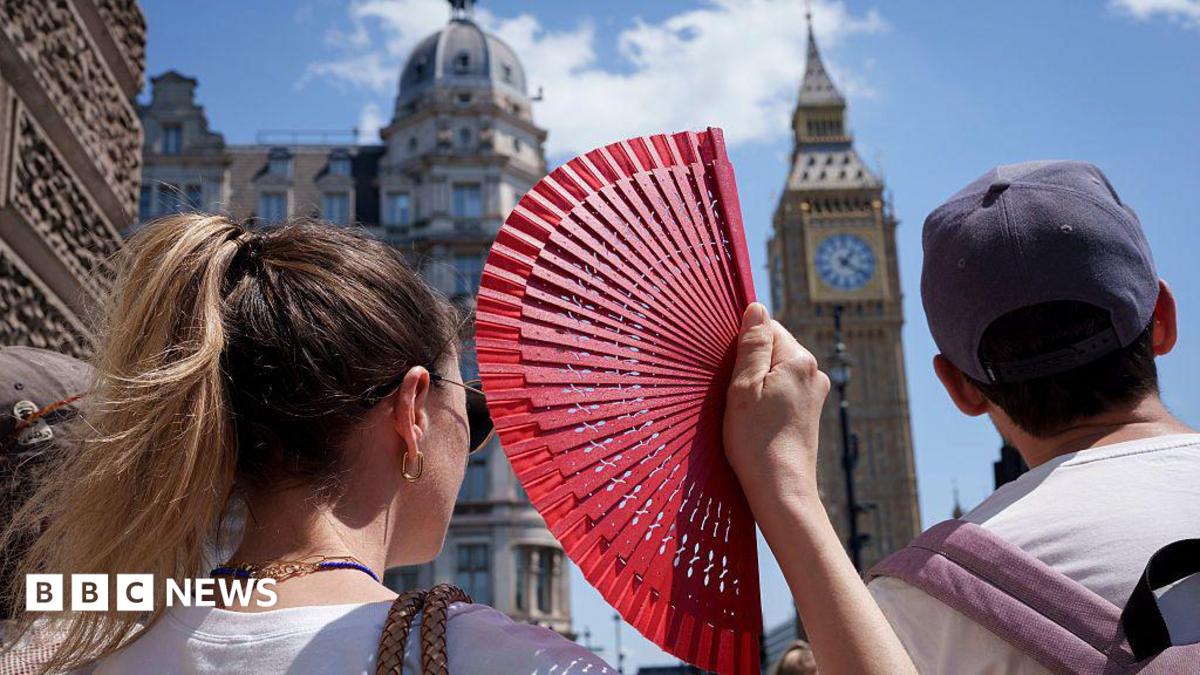 A close-up, over-the-shoulder shot from behind a woman with blonde hair in a ponytail holding up an open red hand fan. Next to her, a man wears a grey baseball cap. In the blurred background, London's Elizabeth clock tower rises against a clear blue sky on a bright, sunny day.