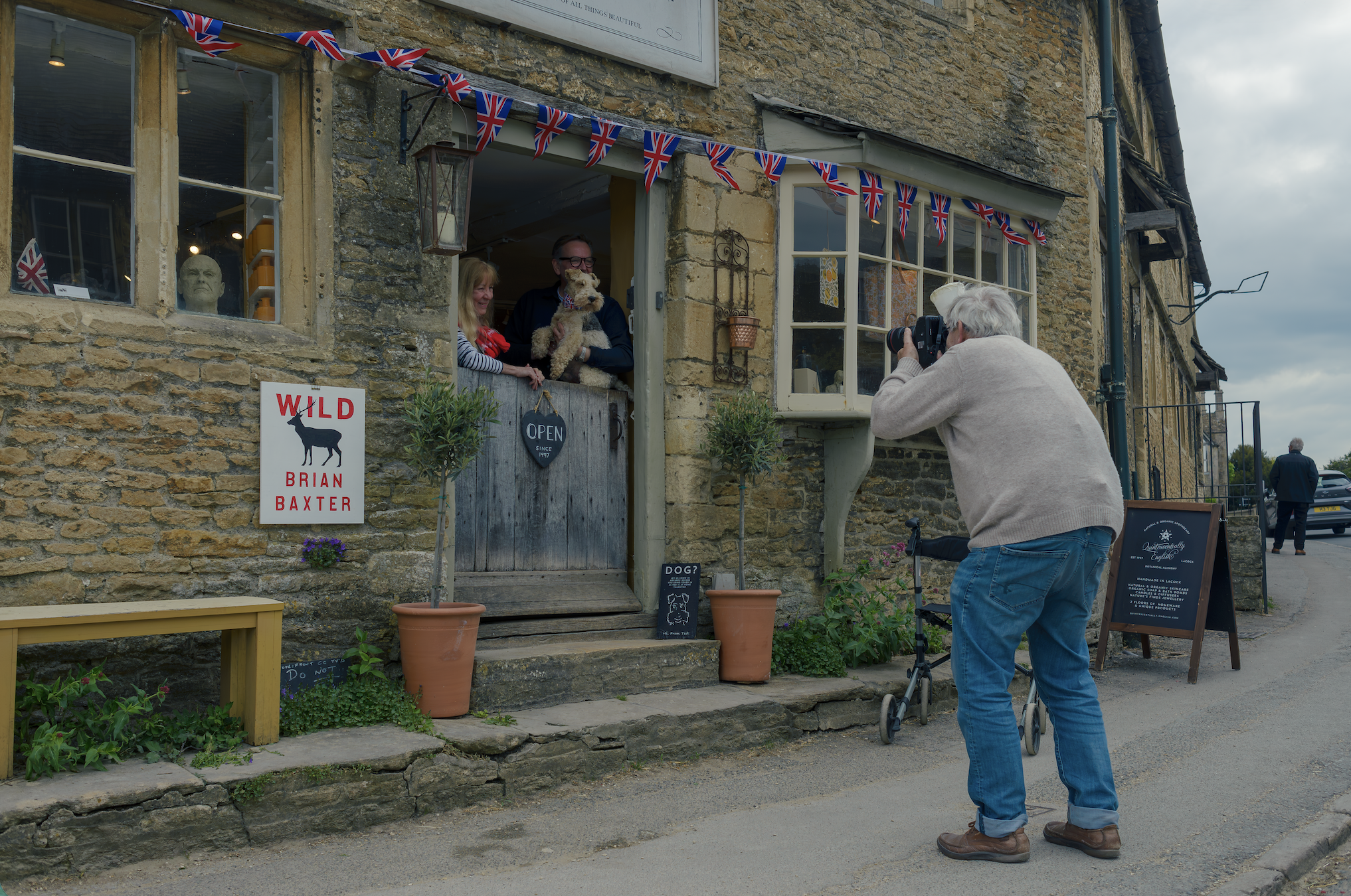 Martin Parr, walking frame beside him, photographs a smiling couple holding a small dog in the doorway of a Union Jack-decorated shop.