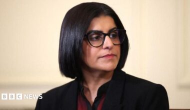 Home Secretary Shabana Mahmood is pictured looking off camera. She is wearing a dark jacket with a red and black top on beneath.