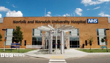 The refurbished outpatients' entrance at the Norfolk and Norwich University Hospital. It shows a white revolving door to the front, with signage to the left and some trees and bushes outside.