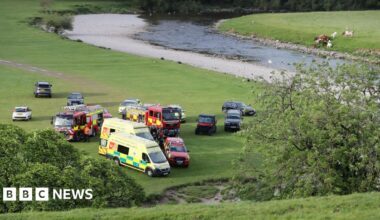 Ambulances, fire engines and other vehicles parked on a trimmed field close to a river meandering nearby.