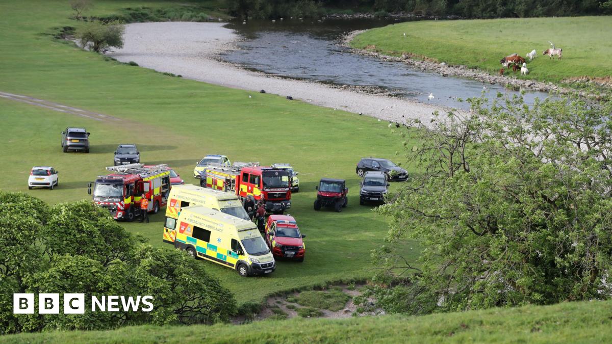 Ambulances, fire engines and other vehicles parked on a trimmed field close to a river meandering nearby.