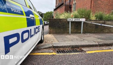 Metropolitan Police car parked beside a street sign reading "Dale Grove N12", with police cordon tape strung across the road and a red-brick church behind.