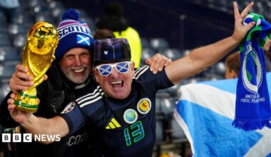 Two smiling Scotland fans during a match in Hampden Stadium, Glasgow. One is holding a replica World Cup and a scarf, and is wearing sunglasses with a saltire pattern. The other has a hat with Scotland written on it.