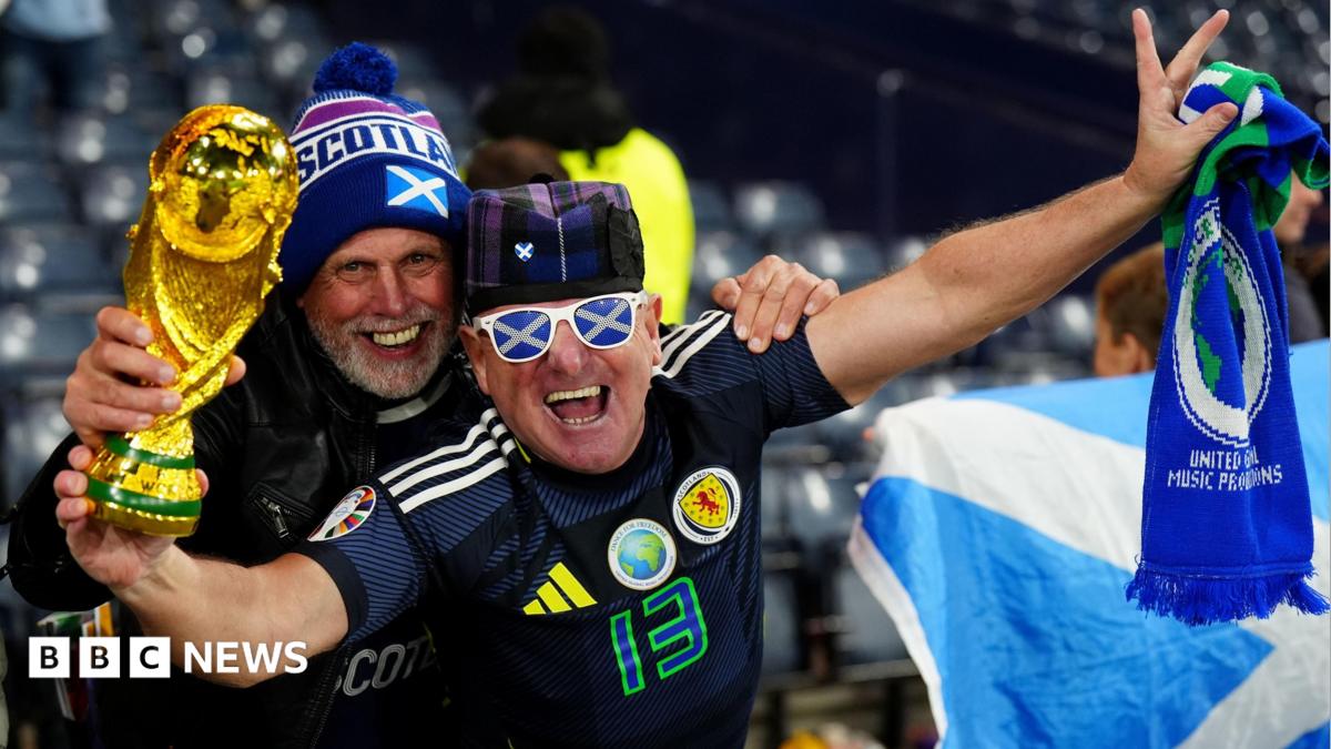 Two smiling Scotland fans during a match in Hampden Stadium, Glasgow. One is holding a replica World Cup and a scarf, and is wearing sunglasses with a saltire pattern. The other has a hat with Scotland written on it.